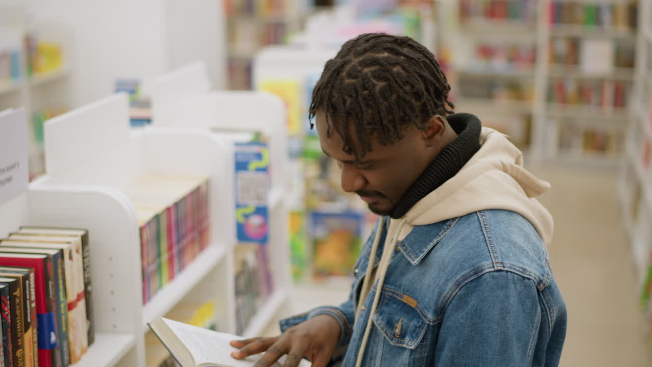 Male in denim jacket examining book in modern library. Focused young man carefully inspects book from shelf in well-organized library, surrounded by colorful books in peaceful, quiet atmosphere