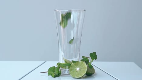 Minzblätter-Fallen-In-Ein-Hohes,-Glänzendes-Glas-Neben-Den-Limettenhälften