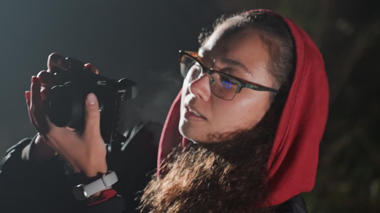 Un fotografo notturno con un cappuccio rosso modifica le impostazioni della fotocamera mentre punta l'obiettivo verso un campo stellato, esaminando il mirino e l'anteprima dell'immagine; radura tranquilla nella foresta con una leggera luceEdge, hobbista concentrato