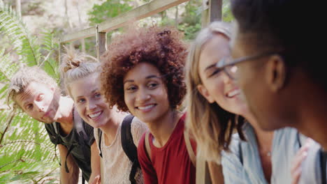 Group-Of-Young-Friends-Hiking-Through-Countryside-Sitting-On-Wooden-Bridge-Together