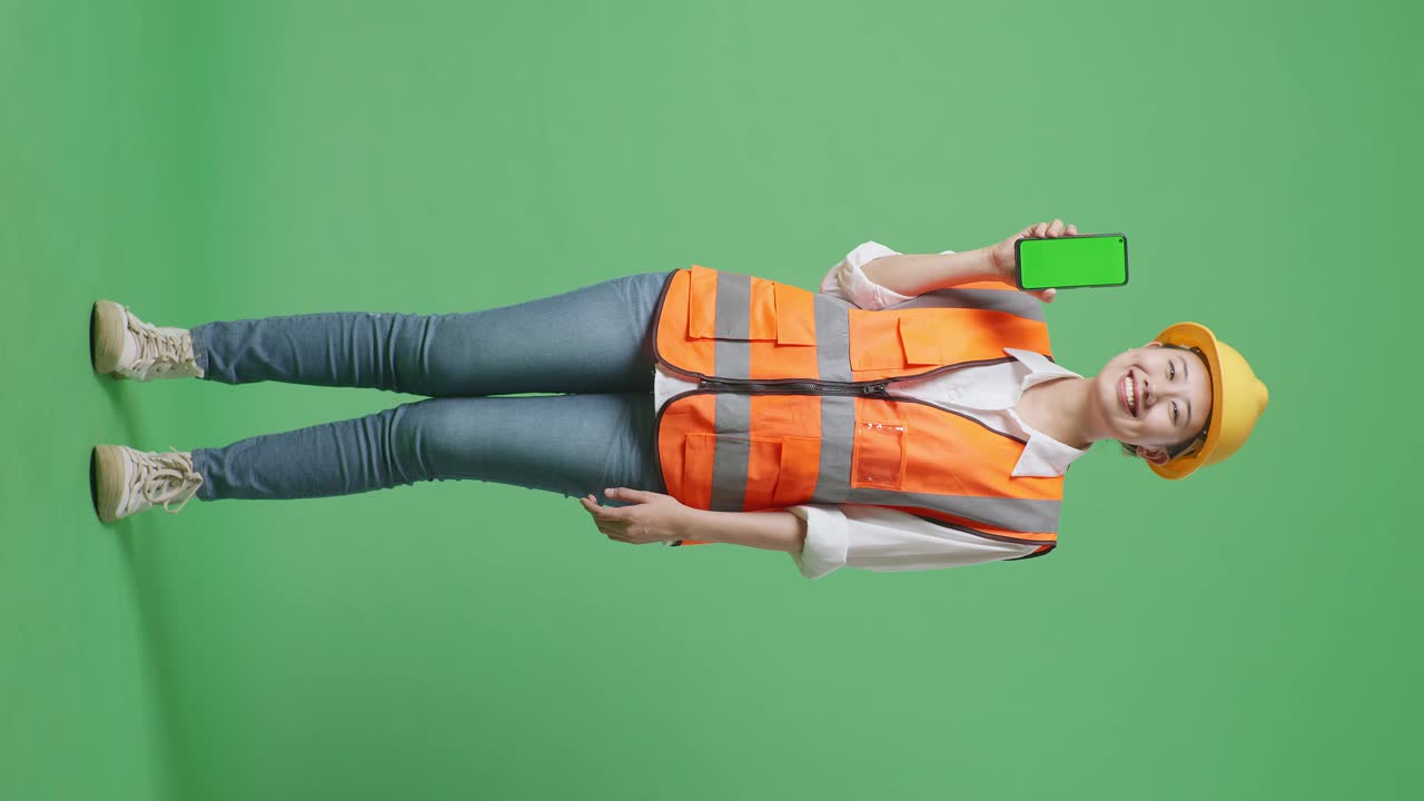 cuerpo lleno de ingeniera asiática con casco de seguridad sonriendo y mostrando teléfono inteligente de pantalla verde a la cámara mientras está de pie en el estudio de fondo de pantalla verde