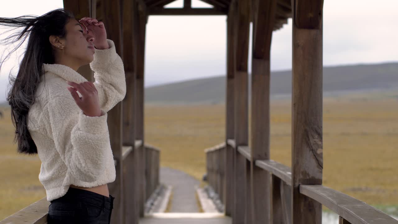 Dark hair woman leans up against wooden bridge rail, fixing hair as it blows in wind