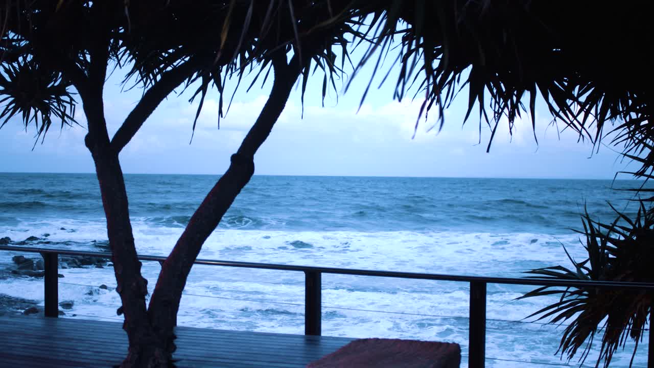 un océano frío y agitado produce pequeñas olas que chocan contra las rocas oscuras que se encuentran debajo de un paseo marítimo de madera, todo iluminado por el sol descendente que emite un tinte azul a todo
