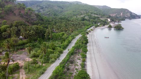 aerial rising above a motorbike travelling along a rough bumpy gravel coastal road surrounded by green trees next to the ocean on idyllic tropical atauro island, timor leste, south east asia