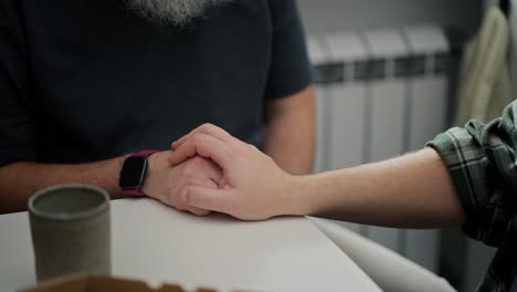 un hombre de mediana edad con una camisa a cuadros toma de la mano a su novio anciano con barba mientras está sentado en una mesa en la cocina mientras habla de su relación como pareja lgbt