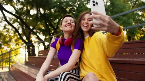 a girl with a short haircut in a purple top and a girl in a yellow sweater take a selfie using a white phone in the park in summer