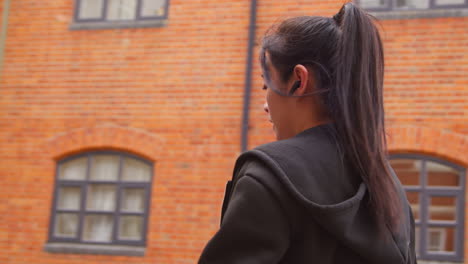 rear view of young woman exercising running along urban street wearing wireless earbuds and smart watch resting