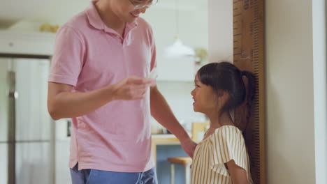 asian father measuring daughter on wall scale at home as she stands on tip toes