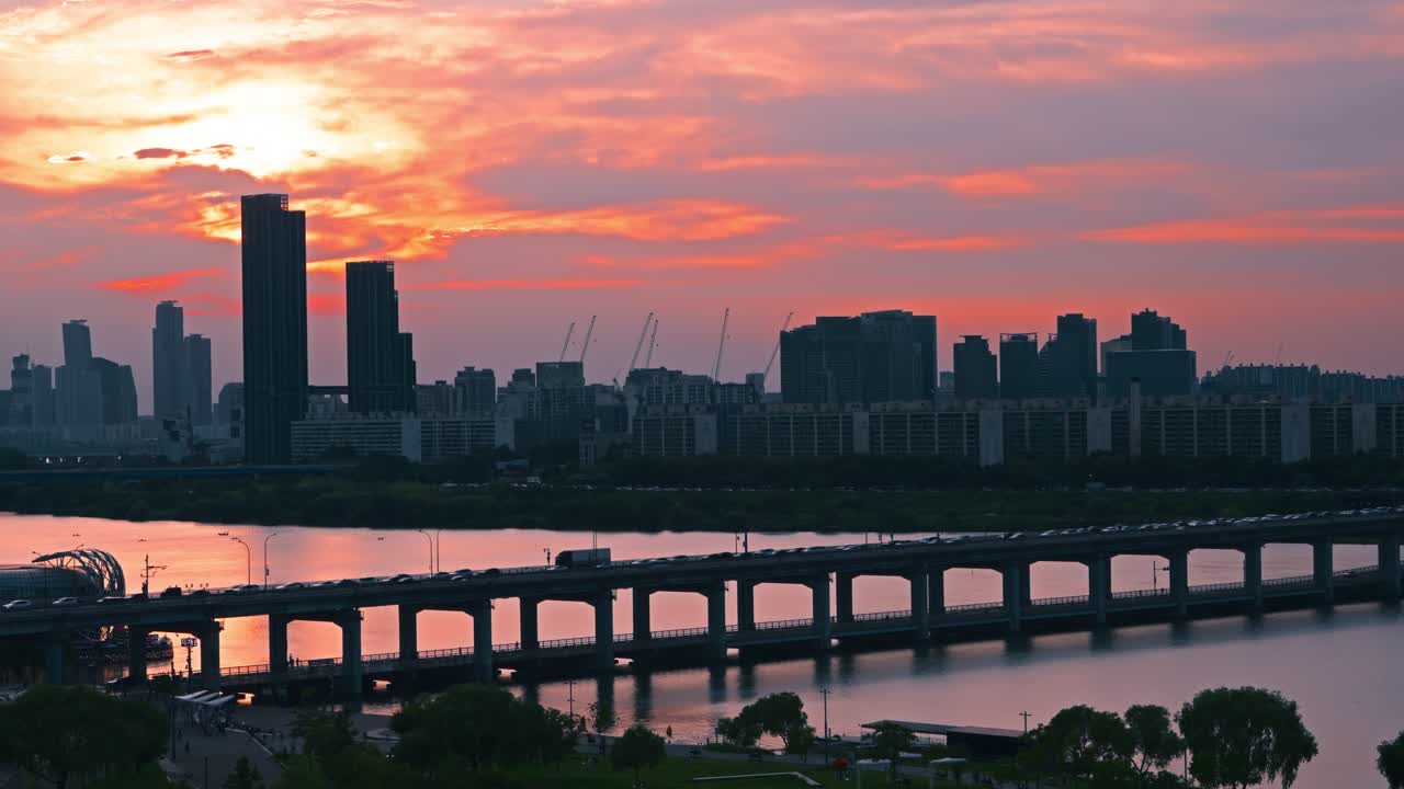 Aerial view of vehicles crossing Banpo Bridge at sunset with dramatic clouds, Han River reflections and city buildings in silhouette