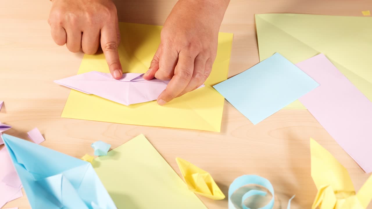 Person folds pastel origami paper on tidy desk, surrounded by finished crafts, under bright lighting