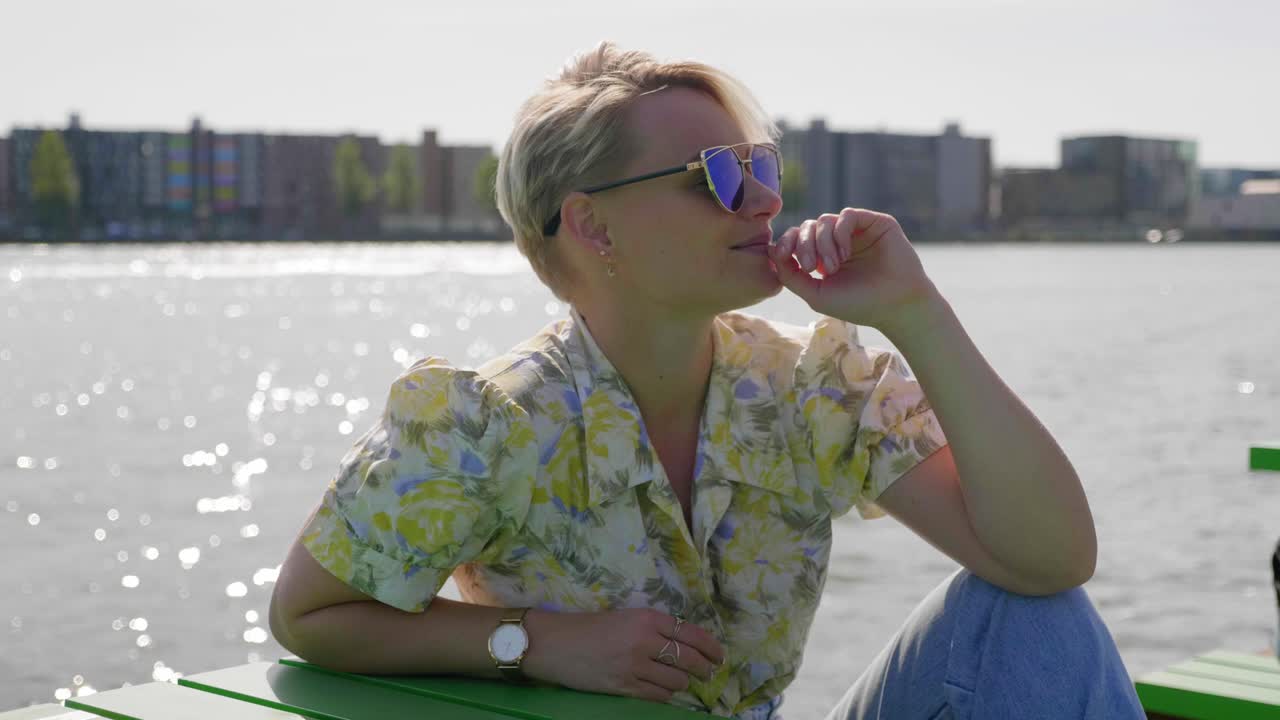 уверенная в себе модная молодая женщина наслаждается теплыми солнечными лучами, сидя у воды