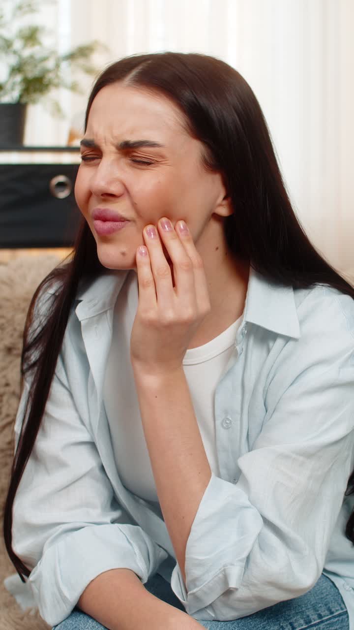 Young woman sitting on sofa holding cheek suffering from strong toothache feeling pain uncomfortable