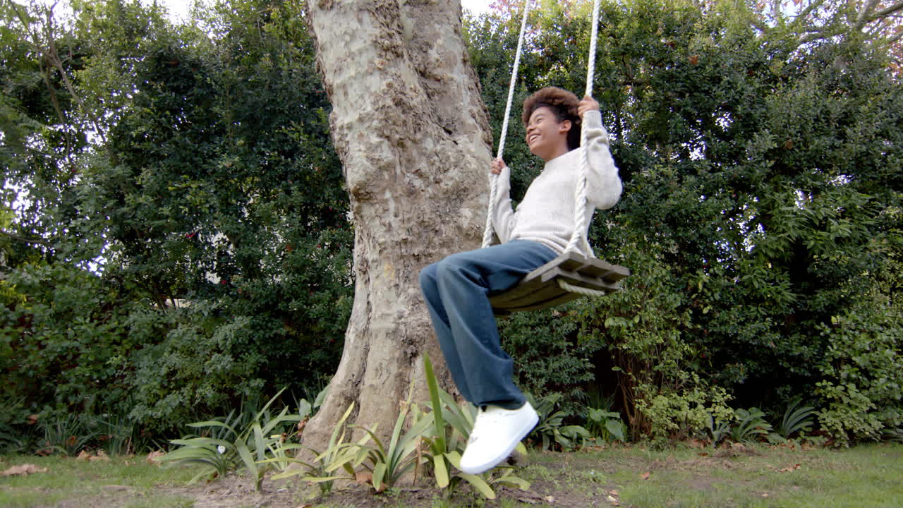 Happy african american boy swinging on tree swing in garden, in slow motion