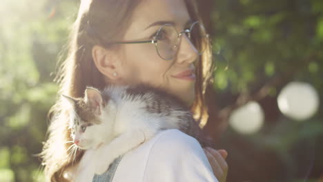 close-up view of a caucasian woman in glasses holding small cat her shoulder in the park on a summer day