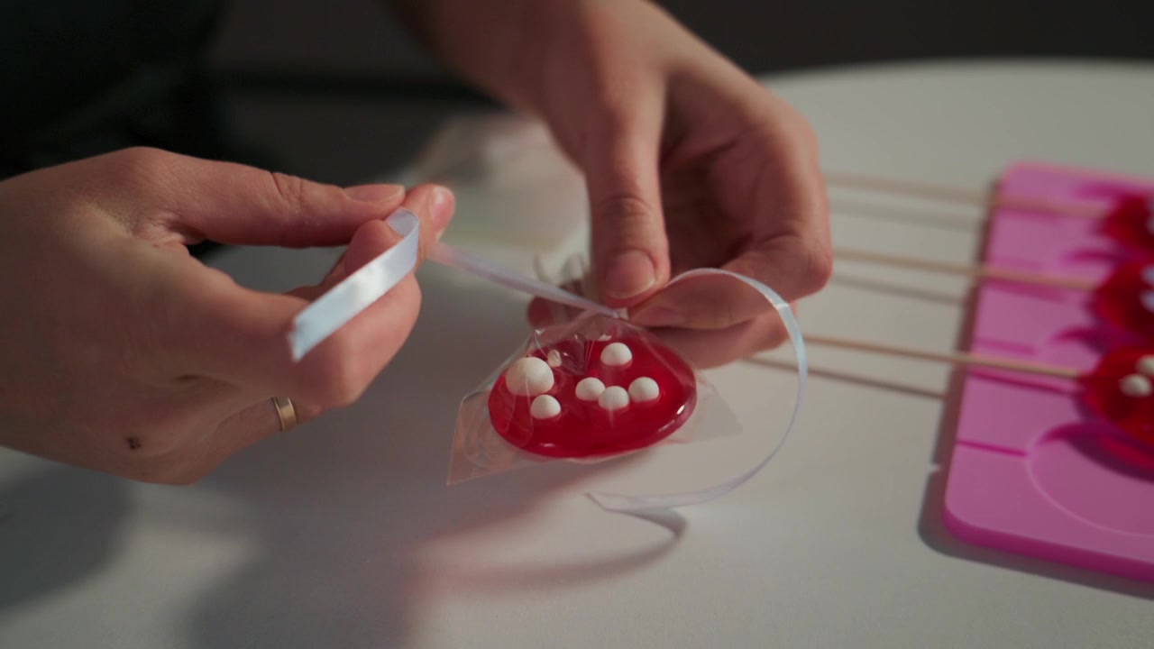 Hands carefully remove glossy red lollipops from a pink mold. Person focuses intently on the task with calm, precise movements. These lollipops feature white sugar pearls. Perfect for food, holiday