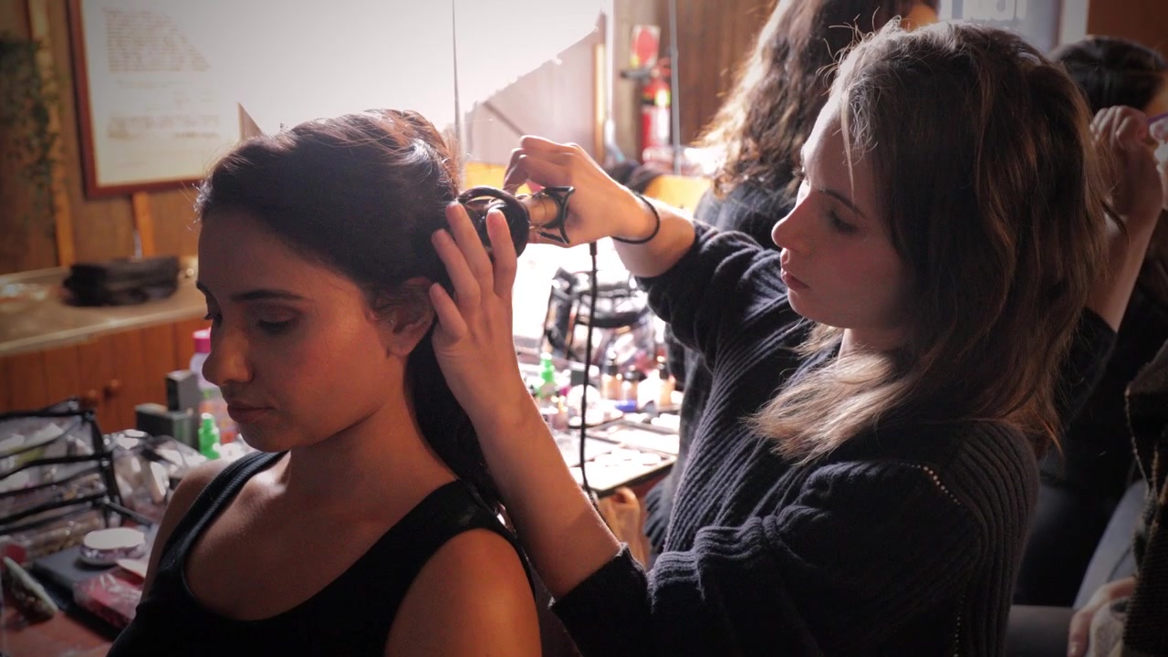 Stylist curls models hair in busy room with several hair and make-up artists preparing for professional photoshoot. HMUA, beautiful models, transformation. Empowerment, concentration and focus. 4K