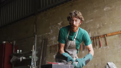 caucasian male blacksmith wearing safety glasses, hammering hot metal tool on anvil in workshop