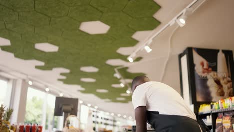 a-Black-skinned-man-in-a-black-apron-lays-out-fruit-and-yakoda-in-special-boxes-on-the-supermarket-counter.-Store-worker-watching-the-products-on-the-counter