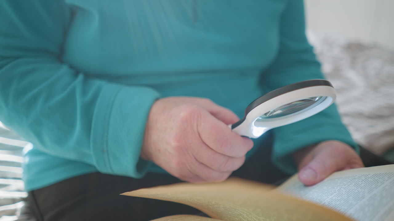 Older woman reading book with magnifying glass. Holding magnifying lens in one hand, looking at page with focused expression. Relaxing in cozy bedroom with natural daylight coming through window