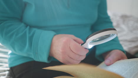 older woman reading book with magnifying glass. holding magnifying lens in one hand, looking at page with focused expression. relaxing in cozy bedroom with natural daylight coming through window