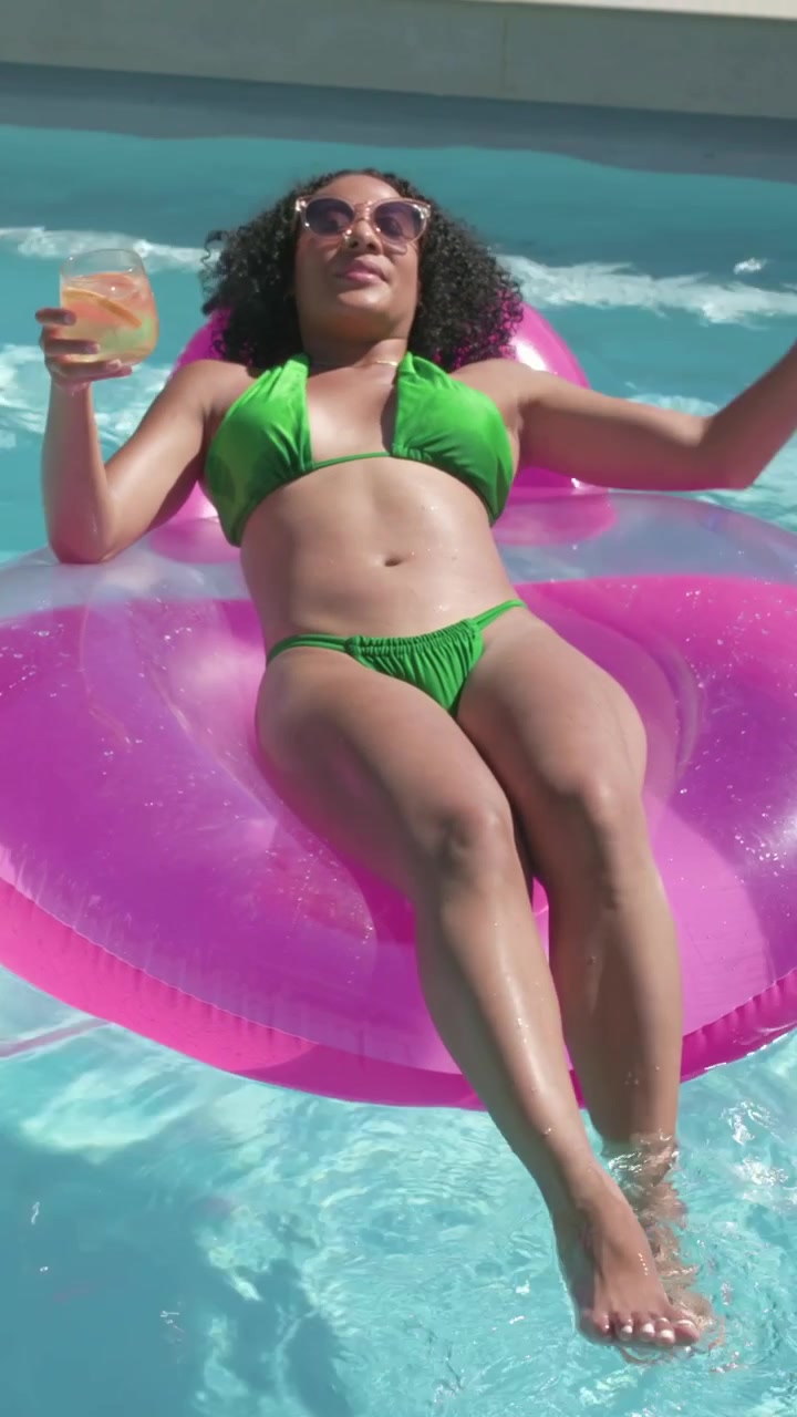 Vertical video: Raising arm woman in green bikini bringing glass into view on pink float, relaxing