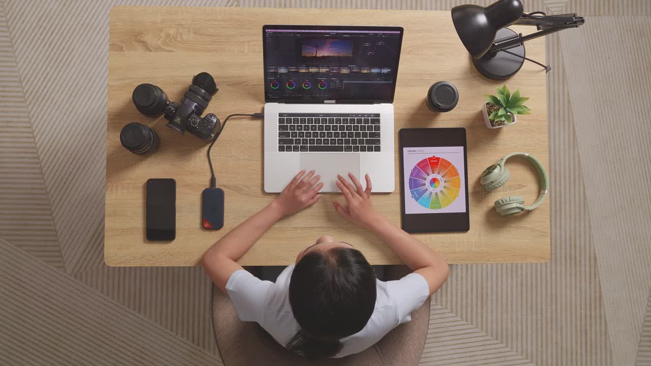 vista superior de una mujer clasificando el color mirando los esquemas de color en una tableta mientras está sentado en el espacio de trabajo usando una computadora portátil al lado de la cámara editando el video en casa