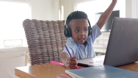 niño afroamericano con auriculares usando una computadora portátil, levantando la mano en una clase en línea en casa, cámara lenta