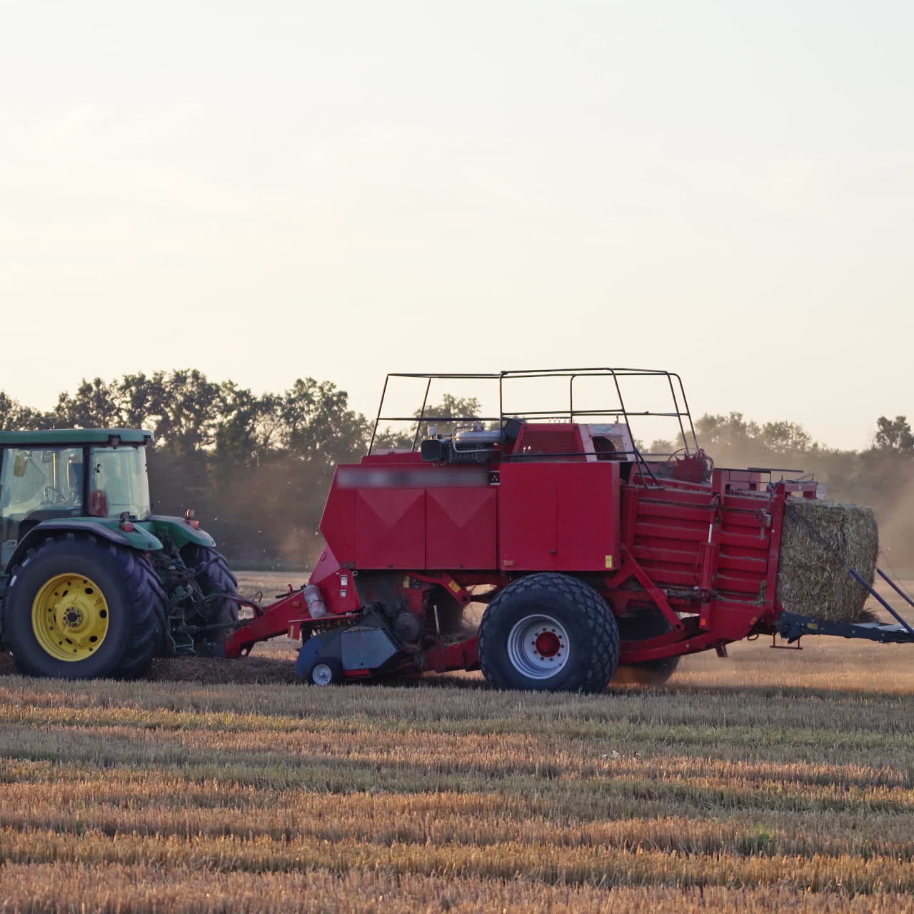 Agricultural machine pressing hay on the golden field. Harvesting dry grass for livestock. Green and red tractor during seasonal works in summer.
