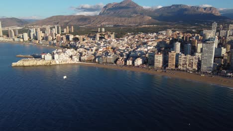 Inclinación-Aérea-Hacia-Arriba-Que-Revela-Los-Edificios-Frente-Al-Mar-De-Benidorm-Con-La-Cordillera-De-Serra-Gelada-En-Segundo-Plano-En-El-Día-Soleado