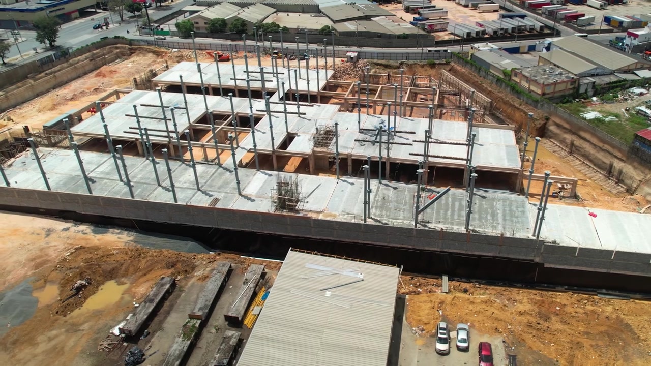 Ripresa aerea con drone ad alta angolazione che si sposta verso destra su un grande cantiere edile commerciale, mostrando pilastri in acciaio, lastre di pavimento in cemento, muro di contenimento e l'area circostante