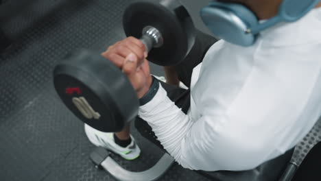 top down view of man in white long sleeve top and black shorts performing dumbbell workout while seated on gym bench, focusing on muscle training and fitness in modern indoor gym environment