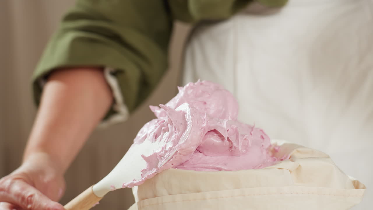 Chef scrapes fluffy pink dough from spatula into container in cozy kitchen, preparing for baking with soft lighting, rustic decor, and baking tools laid out on counter
