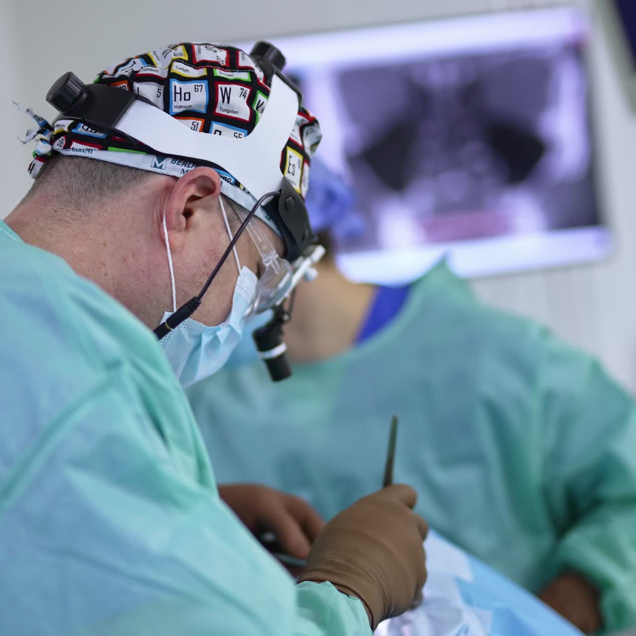 Male surgeon wearing device glasses, cap and mask performs surgery. Doctor standing his side to camera uses tool operating the patient