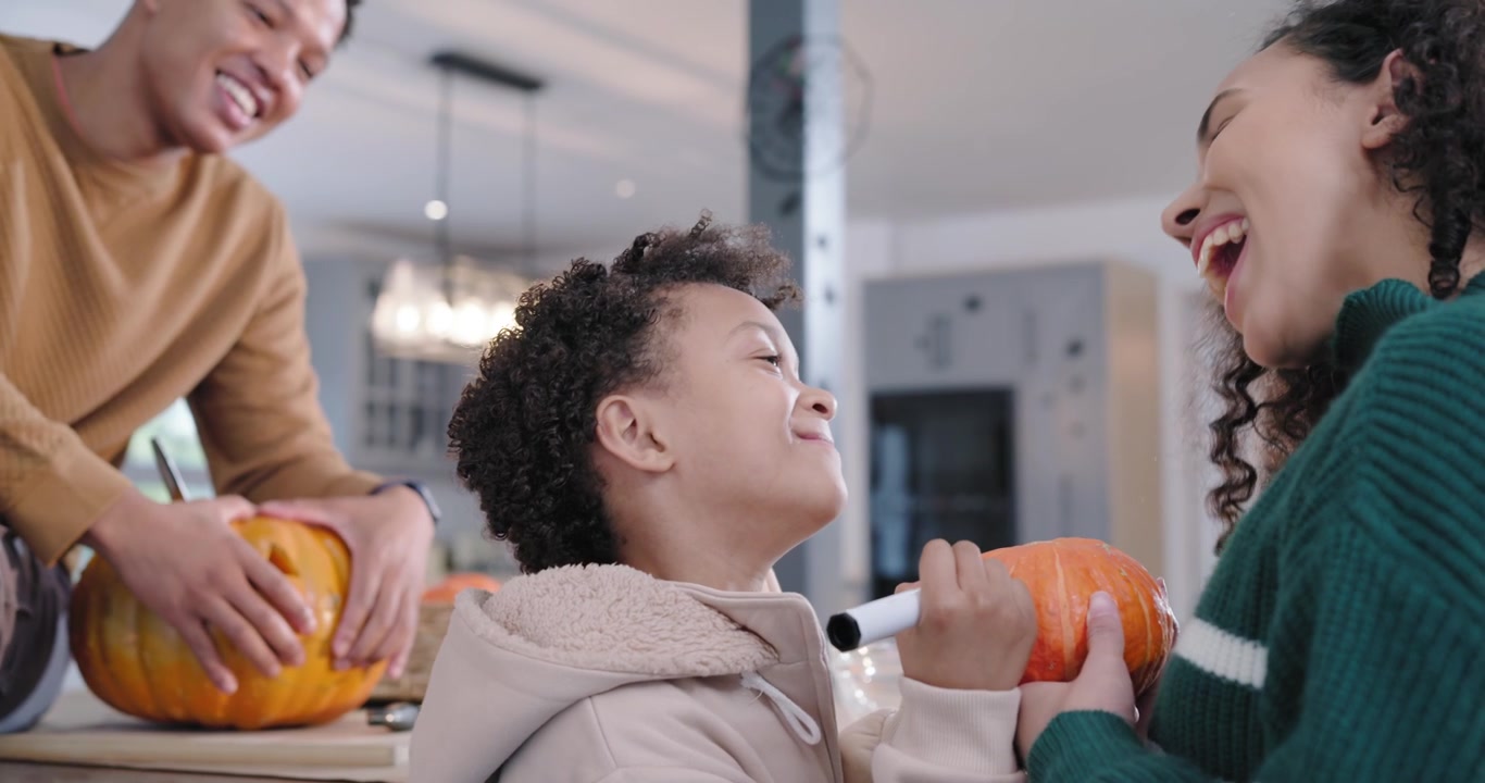 African American family island dad carving pumpkin, mom guiding son decorating with black marker