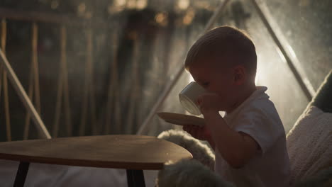 child drinks beverage at low table at glamping. little boy enjoys fresh tea sitting on pillow in night camp. kid with cup and saucer in tent