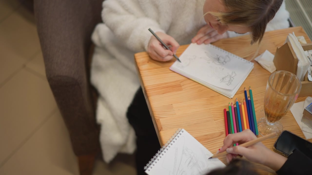 Vista dall'alto mostra due persone sedute a un tavolo di legno che disegnano su quaderni da disegno, con le mani che schizzano attivamente, una matita che si muove sulla pagina mentre un'altra mano offre una matita colorata in un ambiente interno caldo