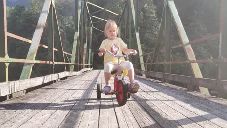 cute blonde child riding tricycle on old wooden bridge - sunlight passing through sidebeams of bridge - handheld moving bakwards while filming girl cycling against camera - eidslandet norway