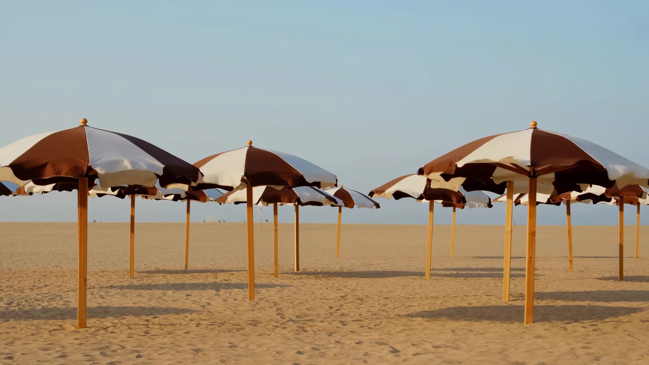 File di ombrelloni da spiaggia a strisce marroni e bianche si ergono da pali di legno su sabbia ampia e aperta, muovendosi dolcemente in una leggera brezza sotto un sole caldo e basso, creando ombre soffuse e un tranquillo ritmo costiero.
