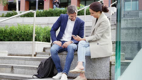 Businessman-And-Businesswoman-Sitting-By-Steps-Having-Meeting-Outdoors-Looking-At-Phone