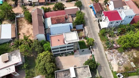 aerial high-angle view of a residential suburb or town in daylight. a narrow road is flanked by varied houses with tiled roofs and green trees, interspersed with open lots.