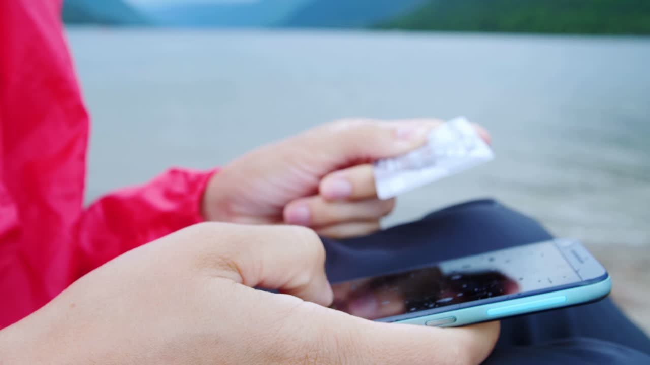 Person using a smartphone and credit card outdoors by a lake