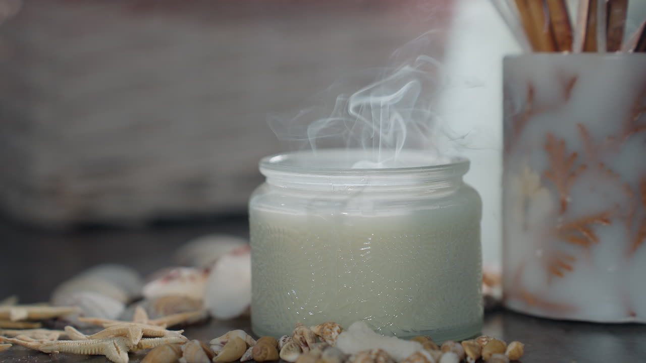 White candle just extinguished releasing gentle smoke on marble table, surrounded by seashells, snail shells, and starfish decor, under soft natural lighting with cozy calm indoor atmosphere