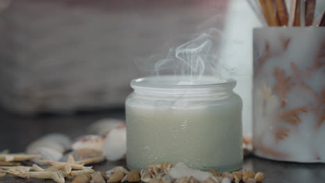 white candle just extinguished releasing gentle smoke on marble table, surrounded by seashells, snail shells, and starfish decor, under soft natural lighting with cozy calm indoor atmosphere