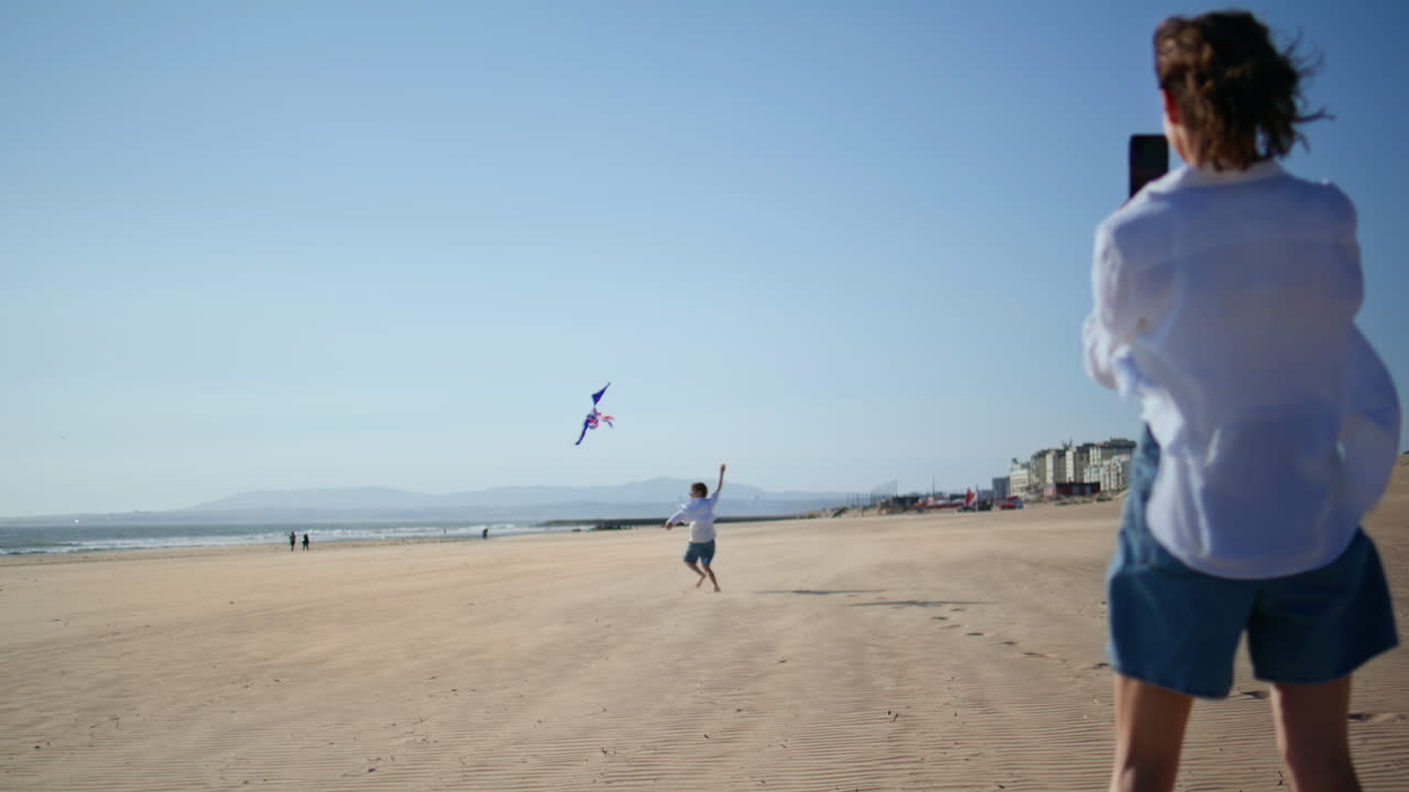 Smiling mother taking picture playing son at smartphone at sunny sandy beach