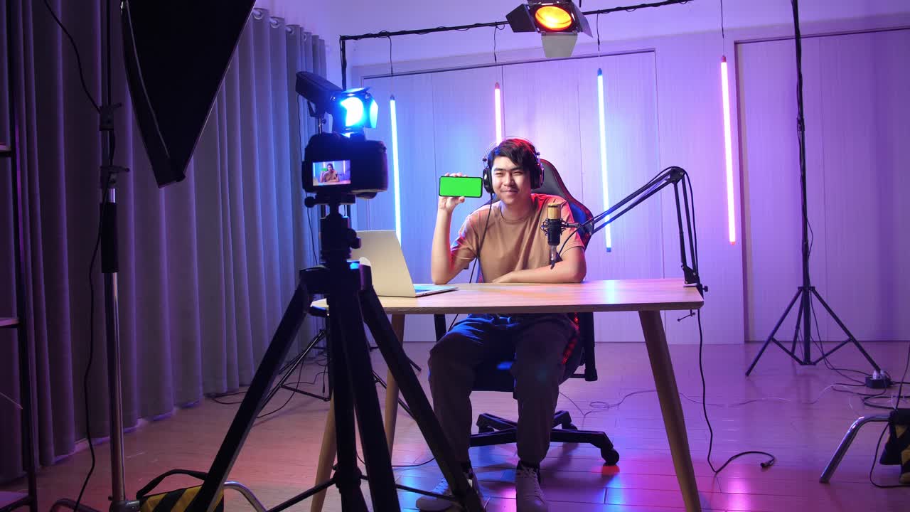 Asian Man Holding Mobile Phone With Mock Up Green Screen While Live Stream With Camera, Professional Light Equipment