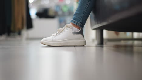 close-up of lady's leg in jeans with white canvas shoes drag her leg back gently tapping left leg gently on floor in well-lit shopping mall