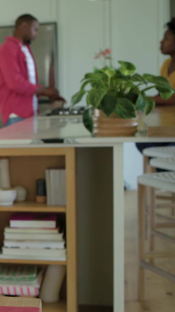 Vertical video: Sitting woman yellow top turning as man entering kitchen using stove to cook, plant