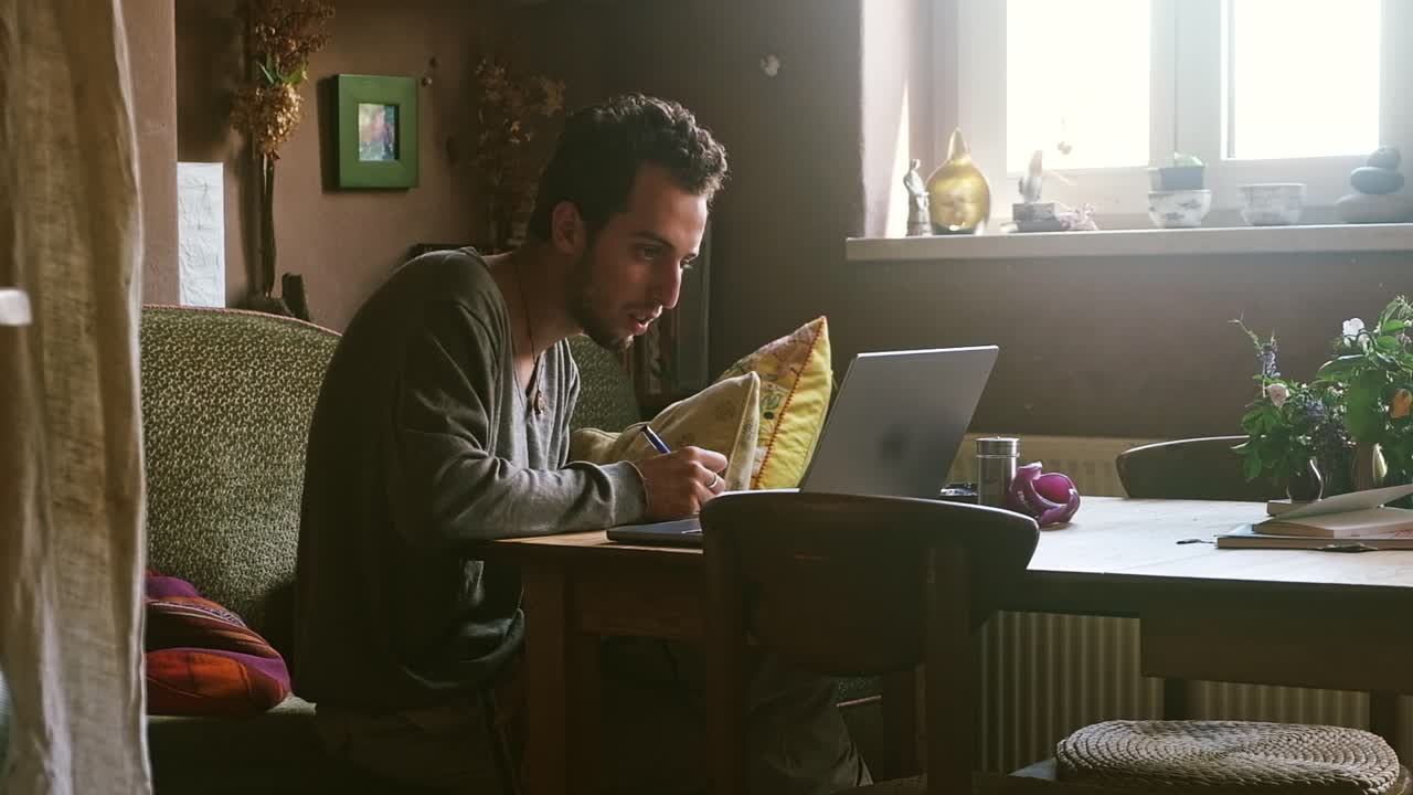 Focused man writing at desk next to laptop in cozy home setting