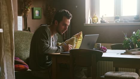 focused man writing at desk next to laptop in cozy home setting