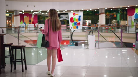 back view of woman in pink dress holding coffee cup and shopping bags while walking in a brightly lit mall looking around, various stores and mall lights are visible, with chair and table by the side